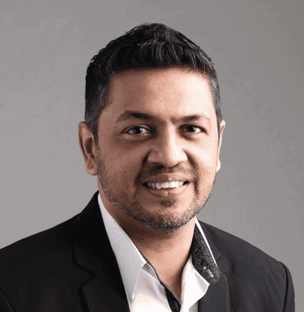 A professional portrait of a smiling man wearing a black blazer over a white shirt, against a light gray background.